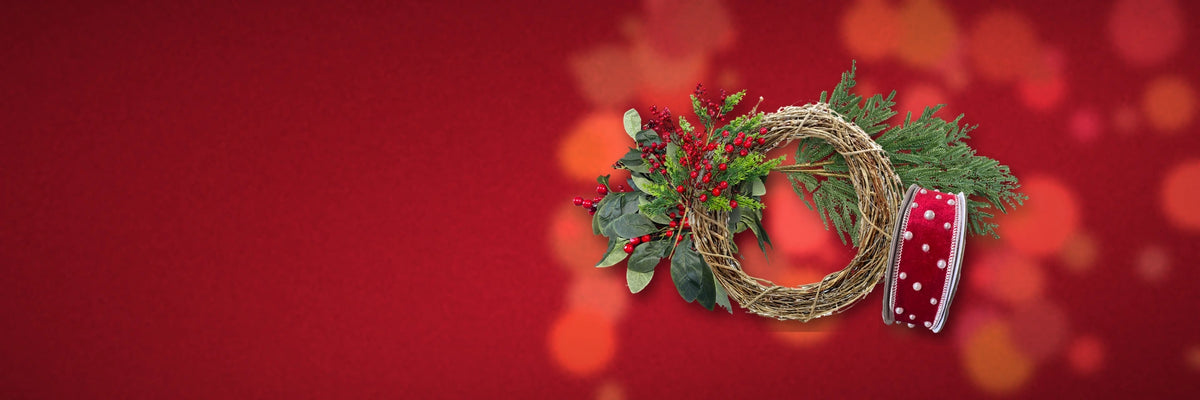 Grapevine wreath base with Christmas pine, red berry sprays and a spool of red velvet ribbon with pearls on a red velvet background.